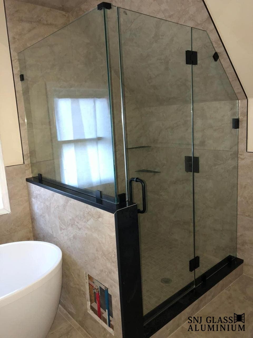 Clear hinged shower door in a gray-tiled bathroom beside a window.