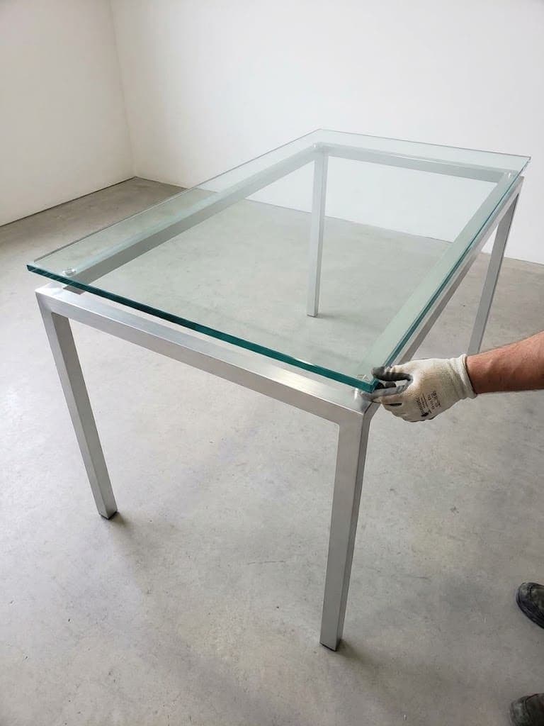 Worker positioning a rectangular clear glass top on a silver metal table frame.