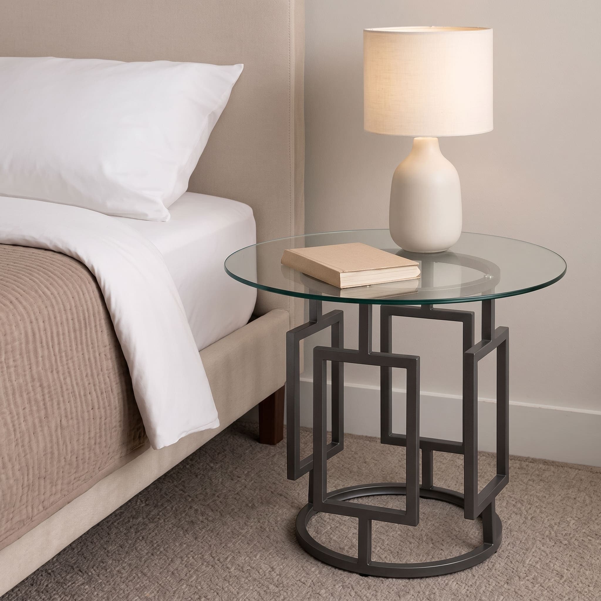Round clear glass bedside table with a dark geometric metal base beside a bed.